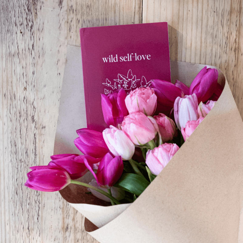 Inspirational Self-Love Book by Lisa Buscomb with pink tulips in a bouquet on a wooden table.