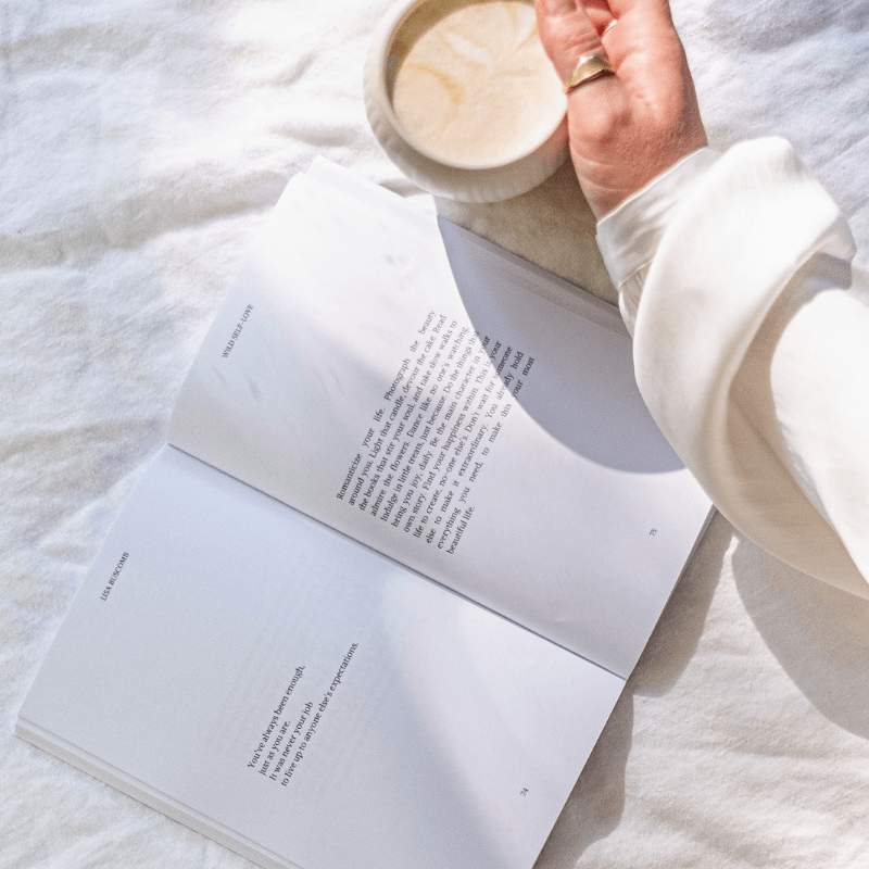 Open pages of the WILD SELF-LOVE Book by Lisa Buscomb with a cup of coffee, showcasing inspirational self-love reminders.