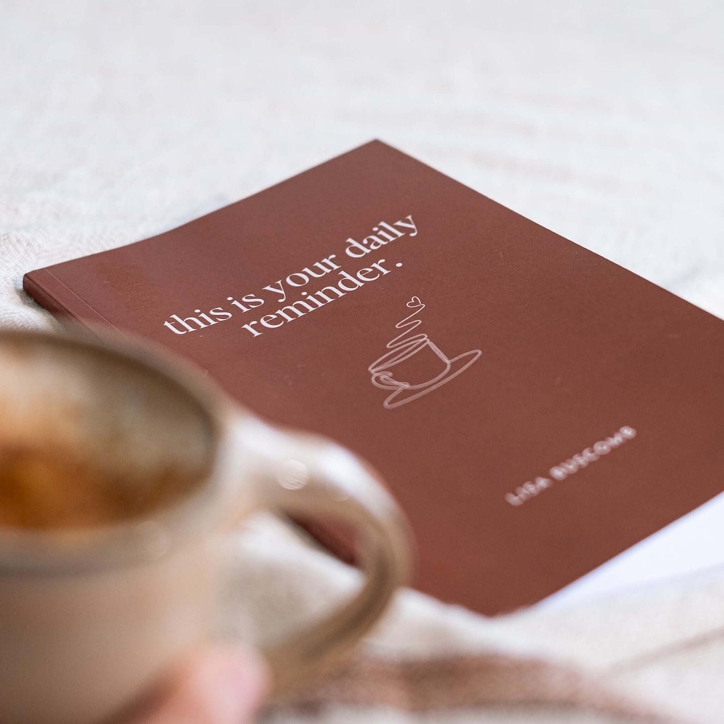 This is Your Daily Reminder Book by Lisa Buscomb resting on a soft surface with a coffee cup nearby.